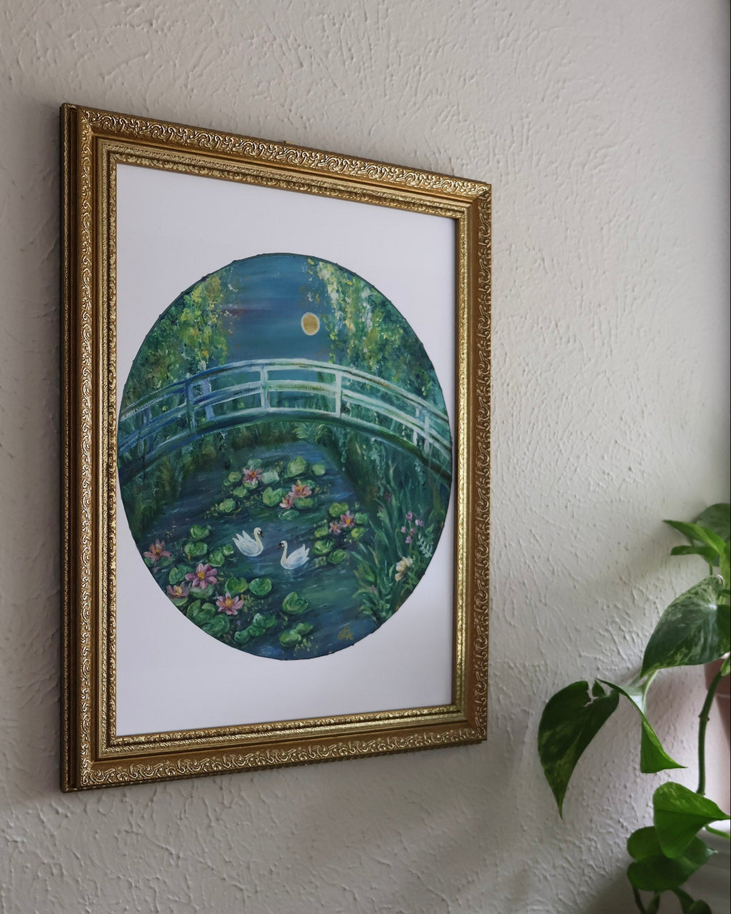 Framed artwork of a bridge over water lilies on a wall with a plant to the right.