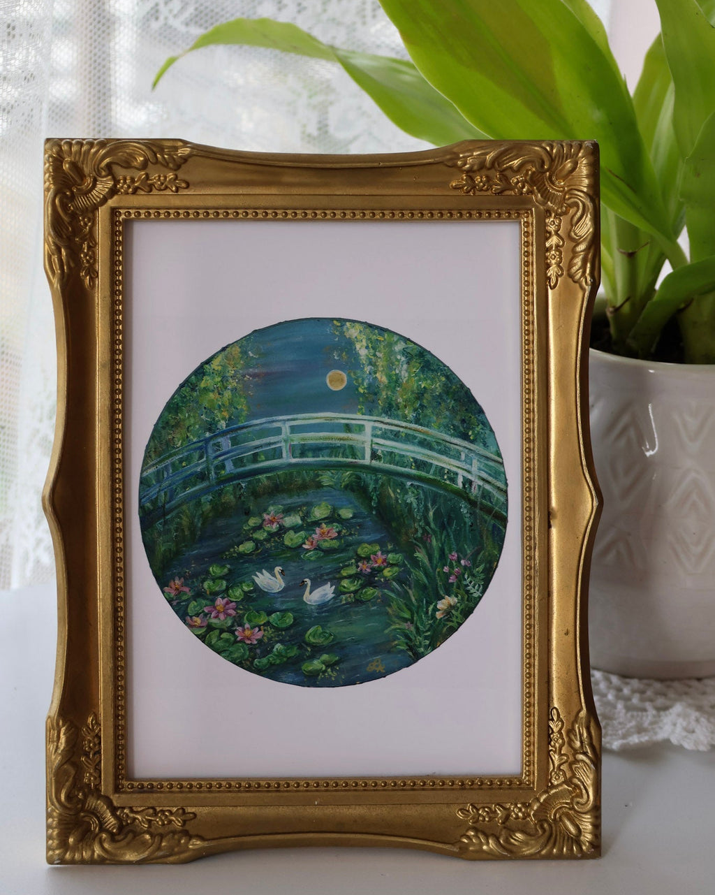 Decorative framed artwork of a garden scene with a gold frame, placed on a surface with a plant in the background.