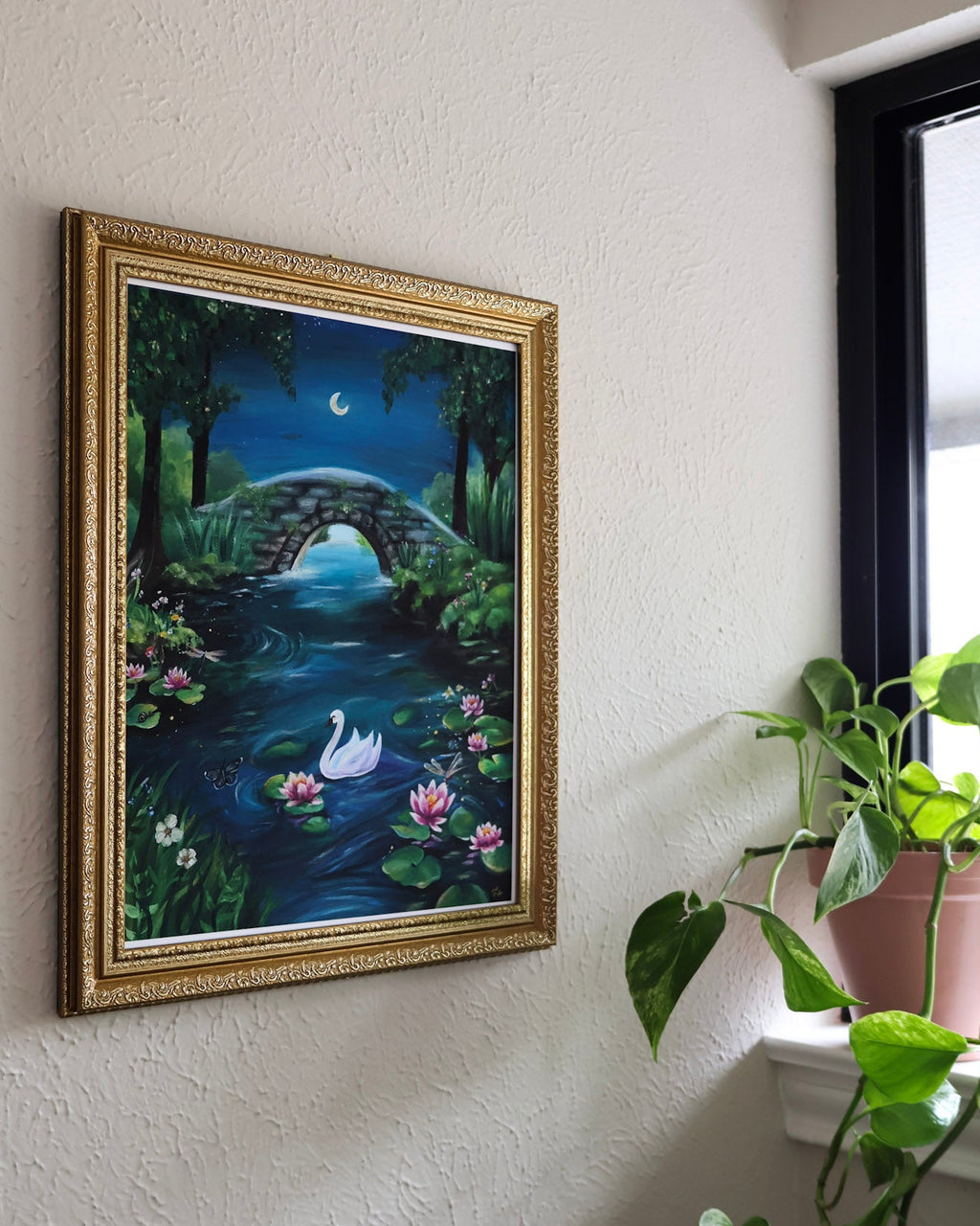 Framed artwork of a nature scene with a swan on a wall next to a window with a plant.
