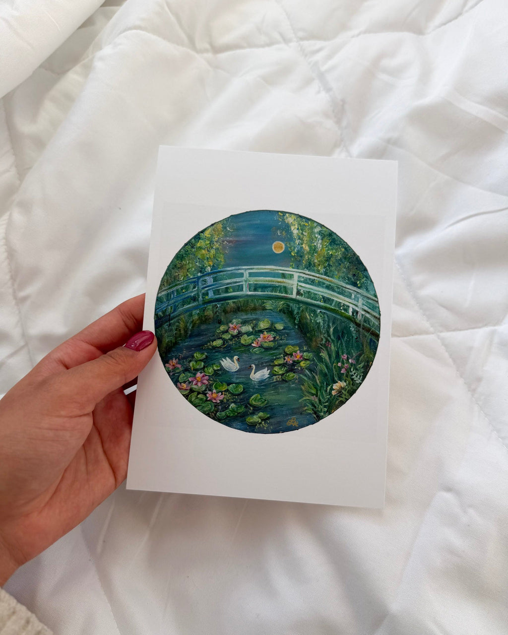 Hand holding a print of a bridge over water lilies against a white quilted background