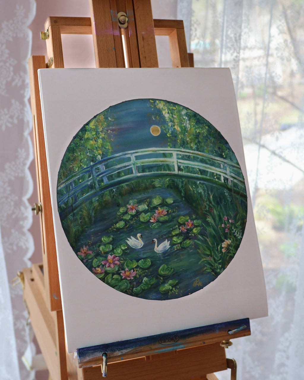 Painting of a bridge over water lilies on an easel with a blurred window background