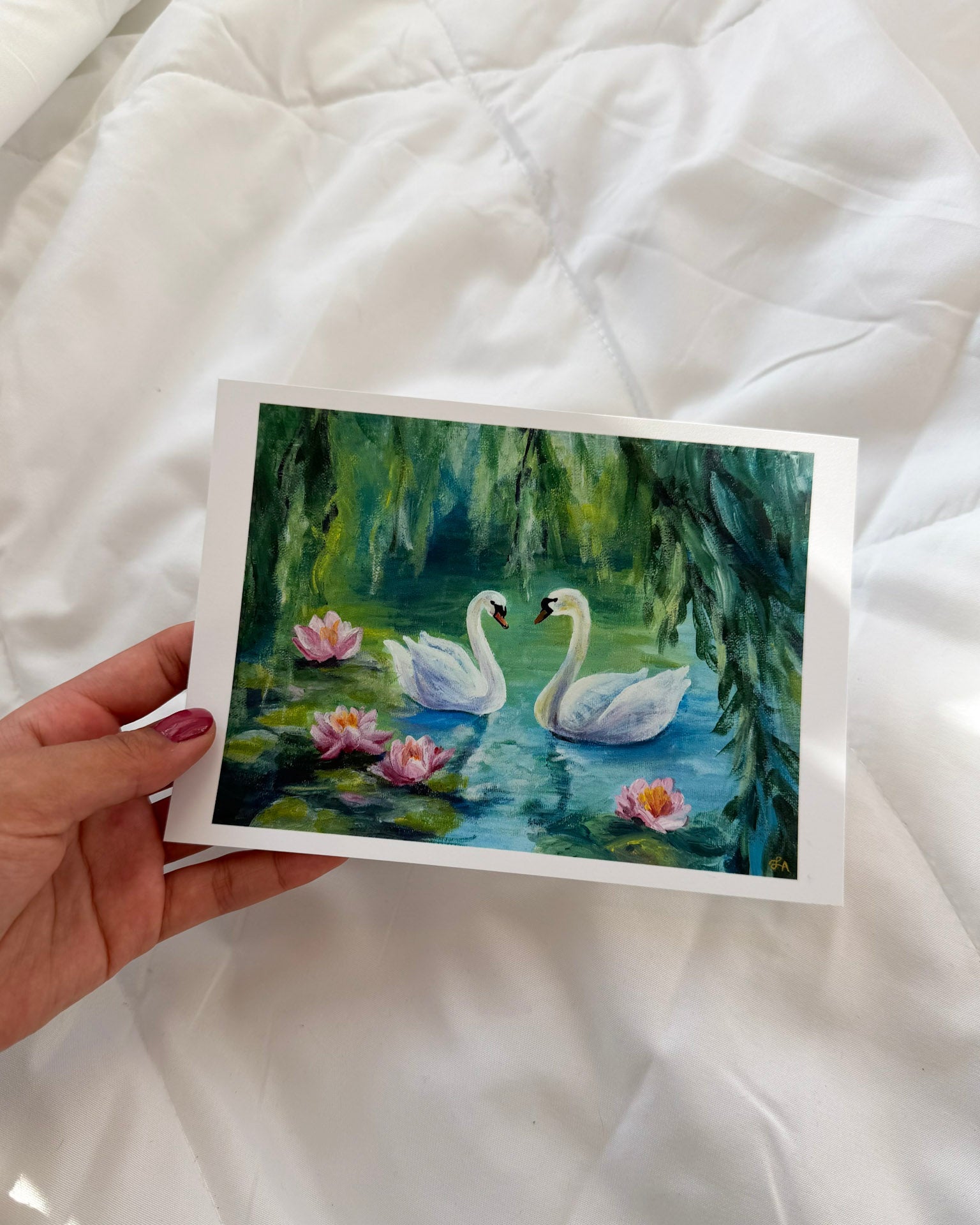 Painting of two swans on water with lilies held by a hand against a white fabric background