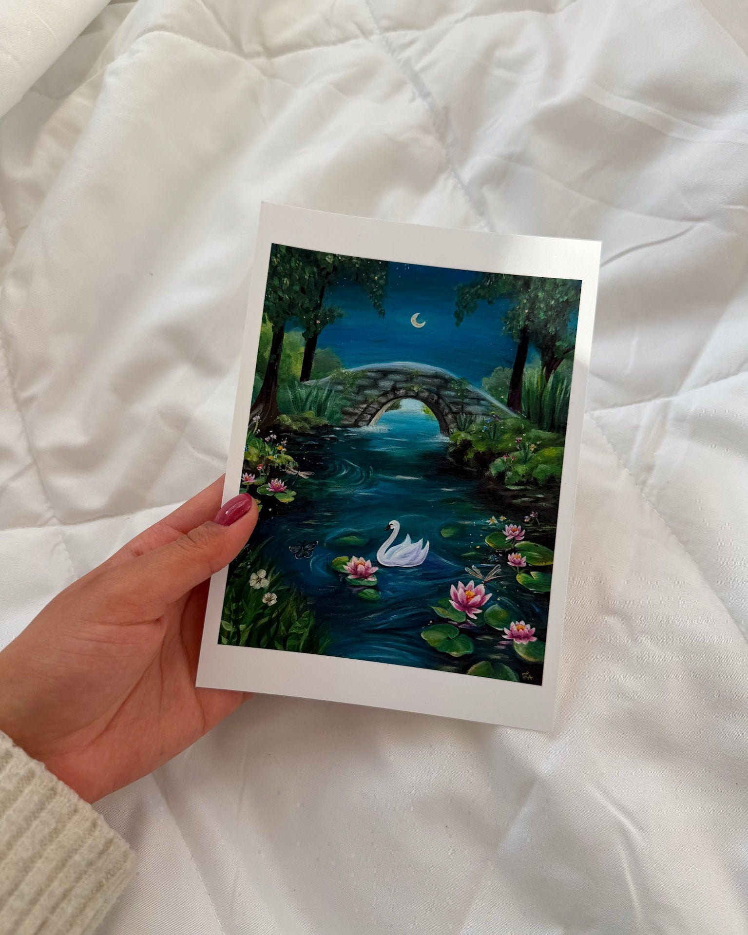 Hand holding a framed artwork of a swan on a pond with lily pads against a white quilted background.