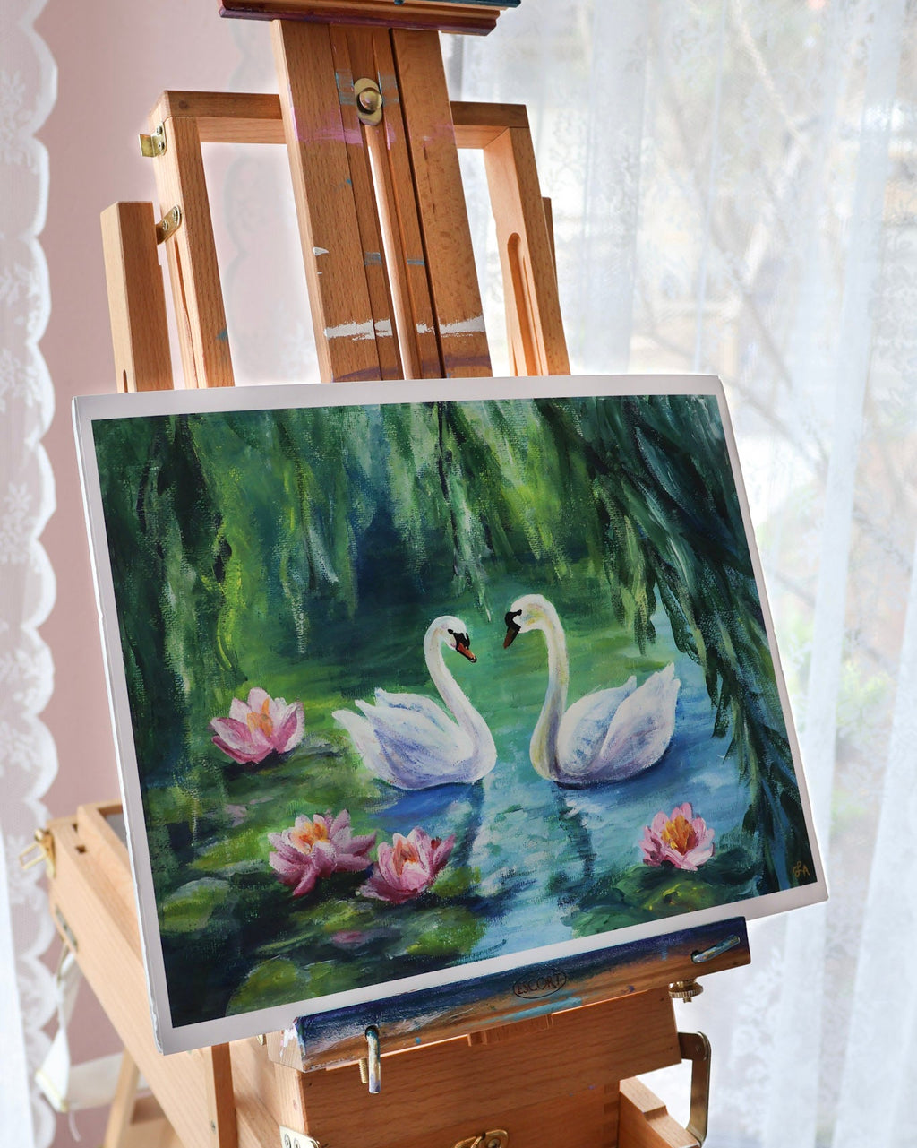 Painting of two swans on a pond with lilies displayed on an easel.