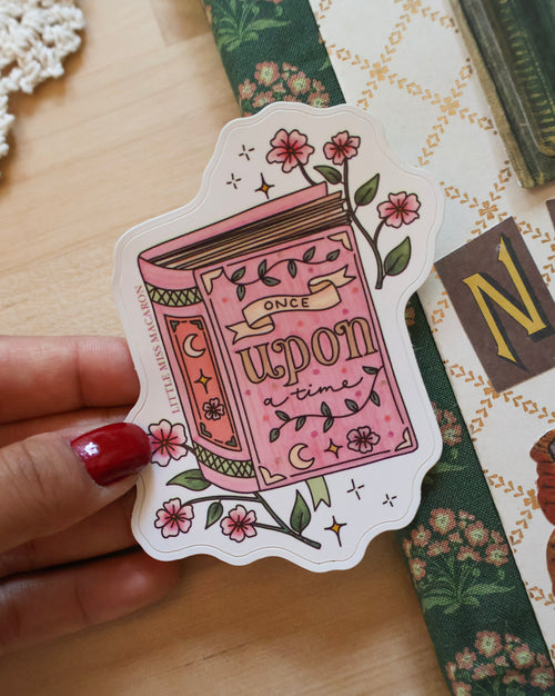 Sticker of a pink book with floral designs held by a hand on a wooden surface.