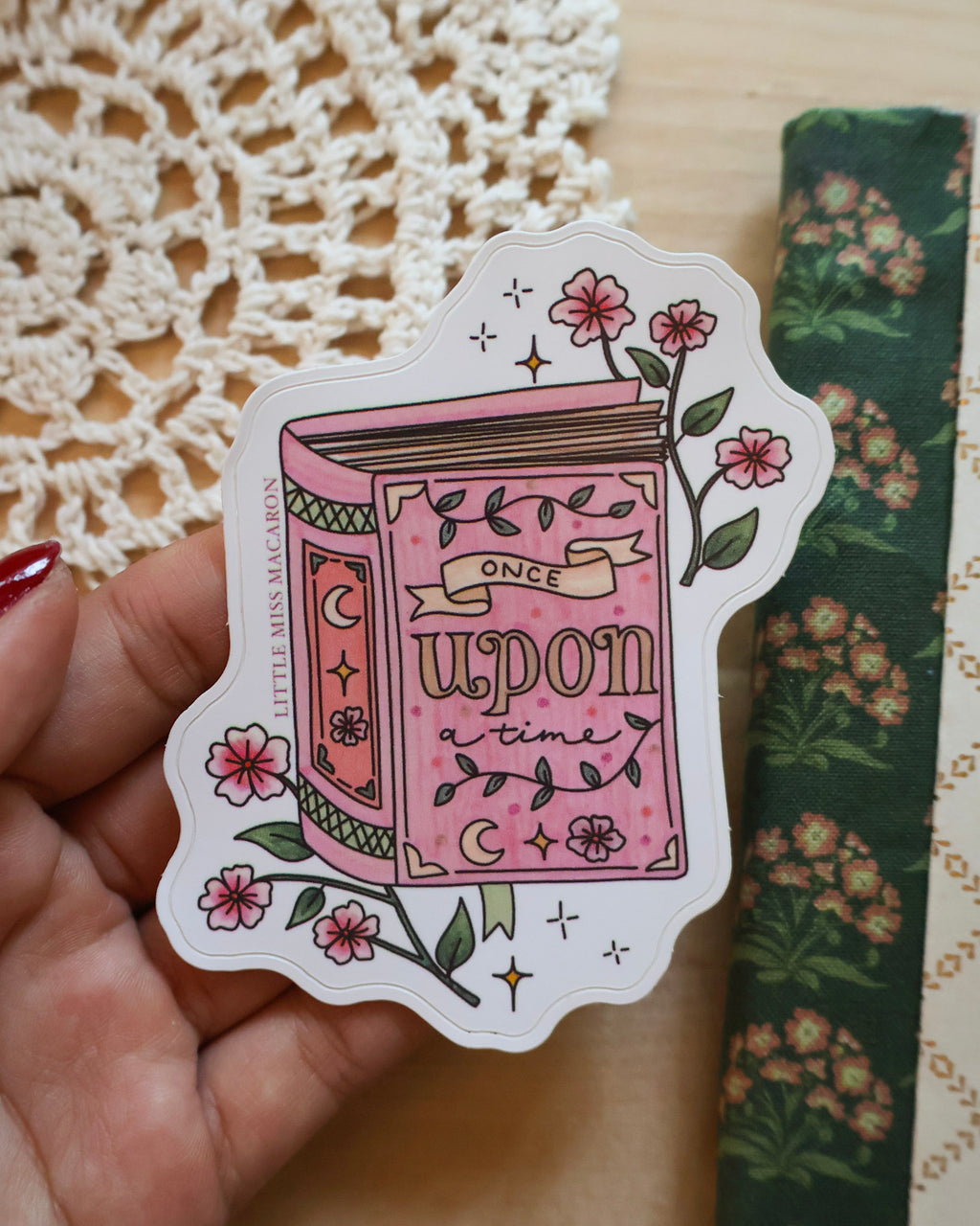 Sticker of a pink book with floral elements held over a textured surface.