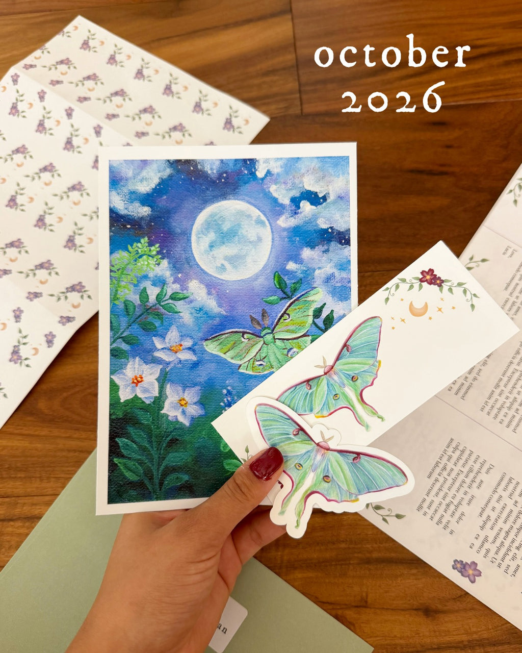 A hand holds a decorative card featuring a blue night sky, moon, and green butterflies, with another butterfly sticker in front. The scene is photographed on a wooden table with patterned paper and an open book around it, and the text 'october 2026' appears in the upper right.