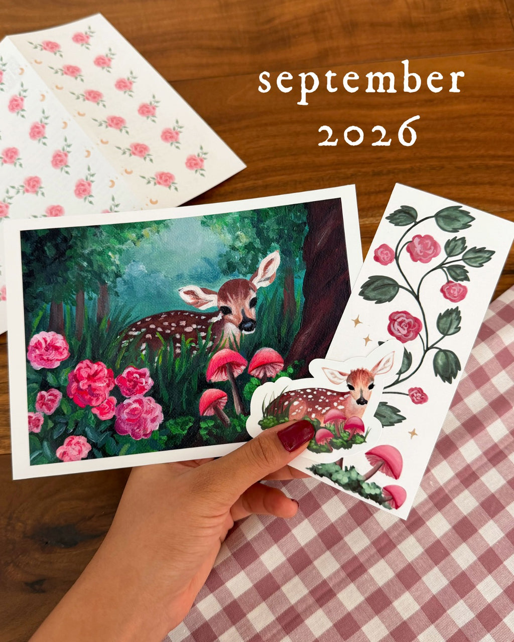 A hand holds printed cards featuring a woodland deer illustration with pink flowers and mushrooms, alongside floral patterned cards, on a wooden surface with a pink-and-white gingham cloth. The text 'september 2026' appears in the upper right.